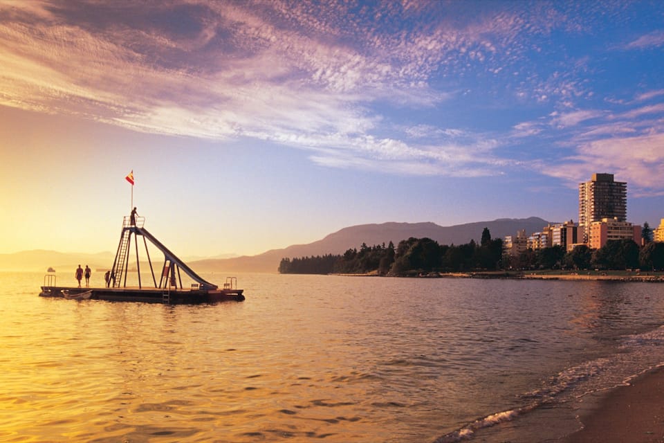 A sunset over a calm body of water with a small boat and people on a dock, a beach, and a cityscape with buildings in the background.