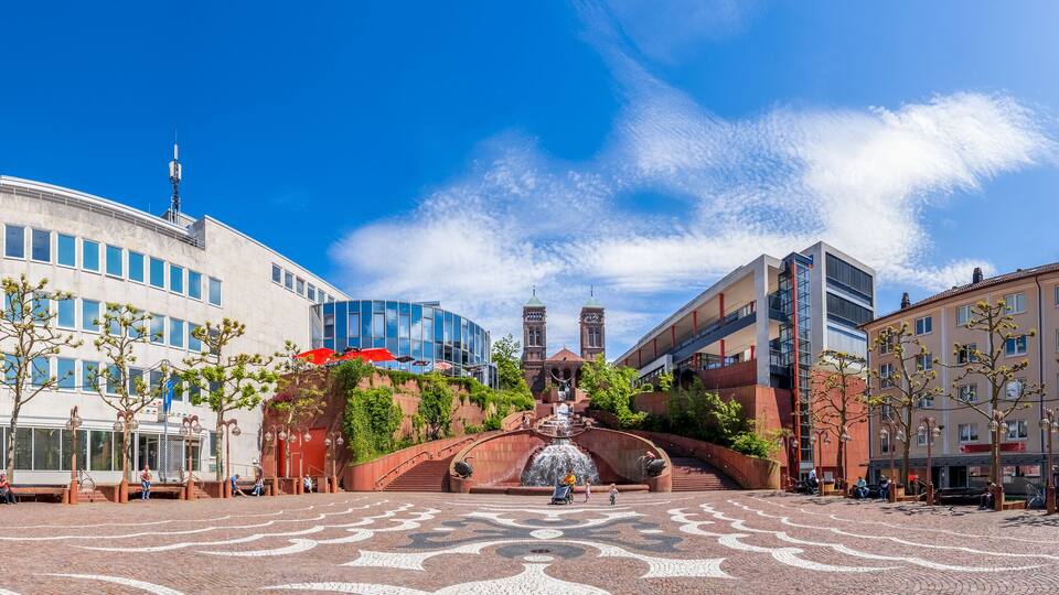 A plaza with a fountain, modern buildings, and cobblestone pavement.