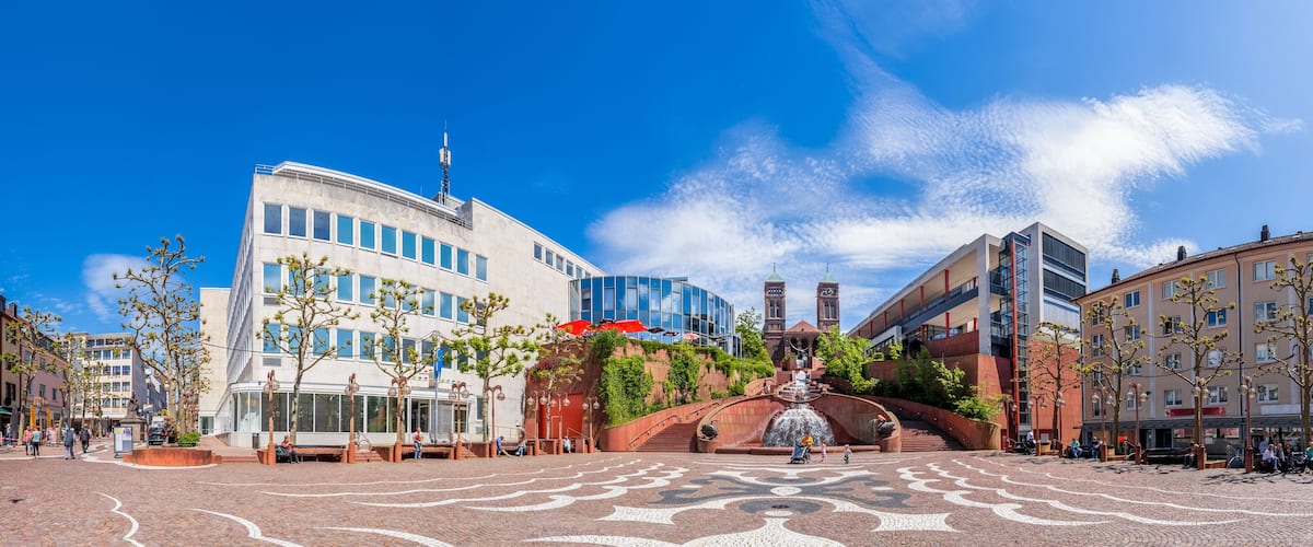 A plaza with a fountain, modern buildings, and cobblestone pavement.