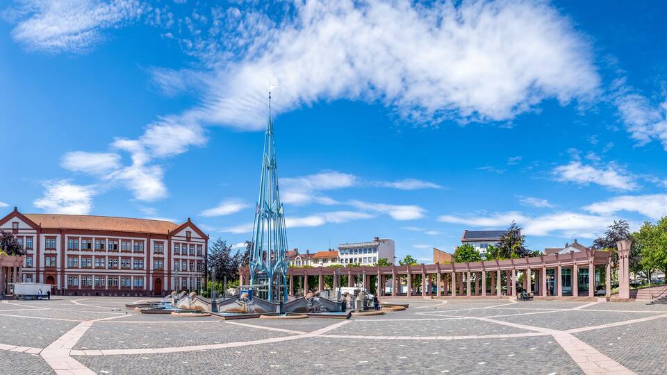 A plaza with a central fountain, surrounding buildings, and a clear blue sky.