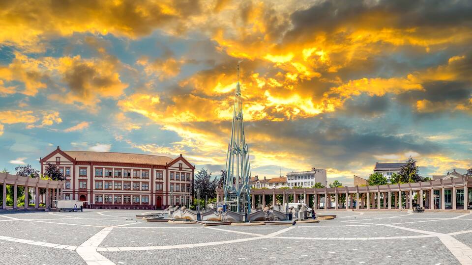 A plaza with a central fountain, surrounded by classical architecture and a dramatic sunset sky.