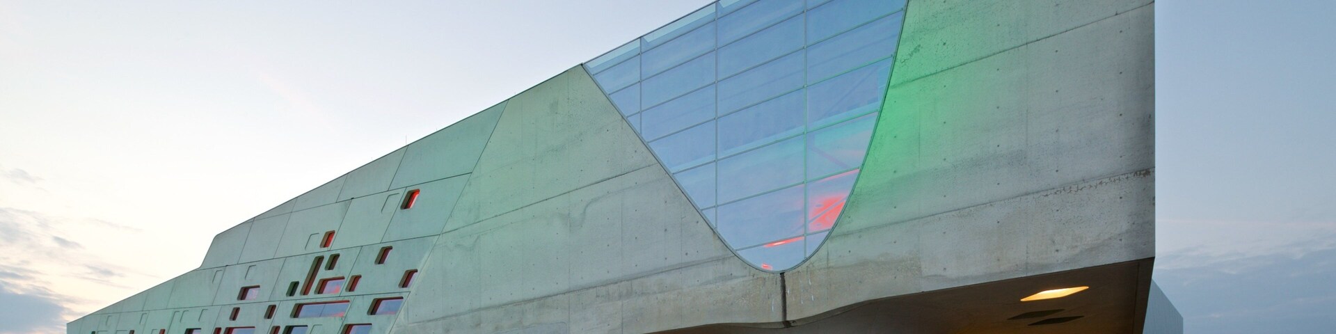 A modern architectural building with a unique angular design, featuring a large glass facade and multiple illuminated windows.