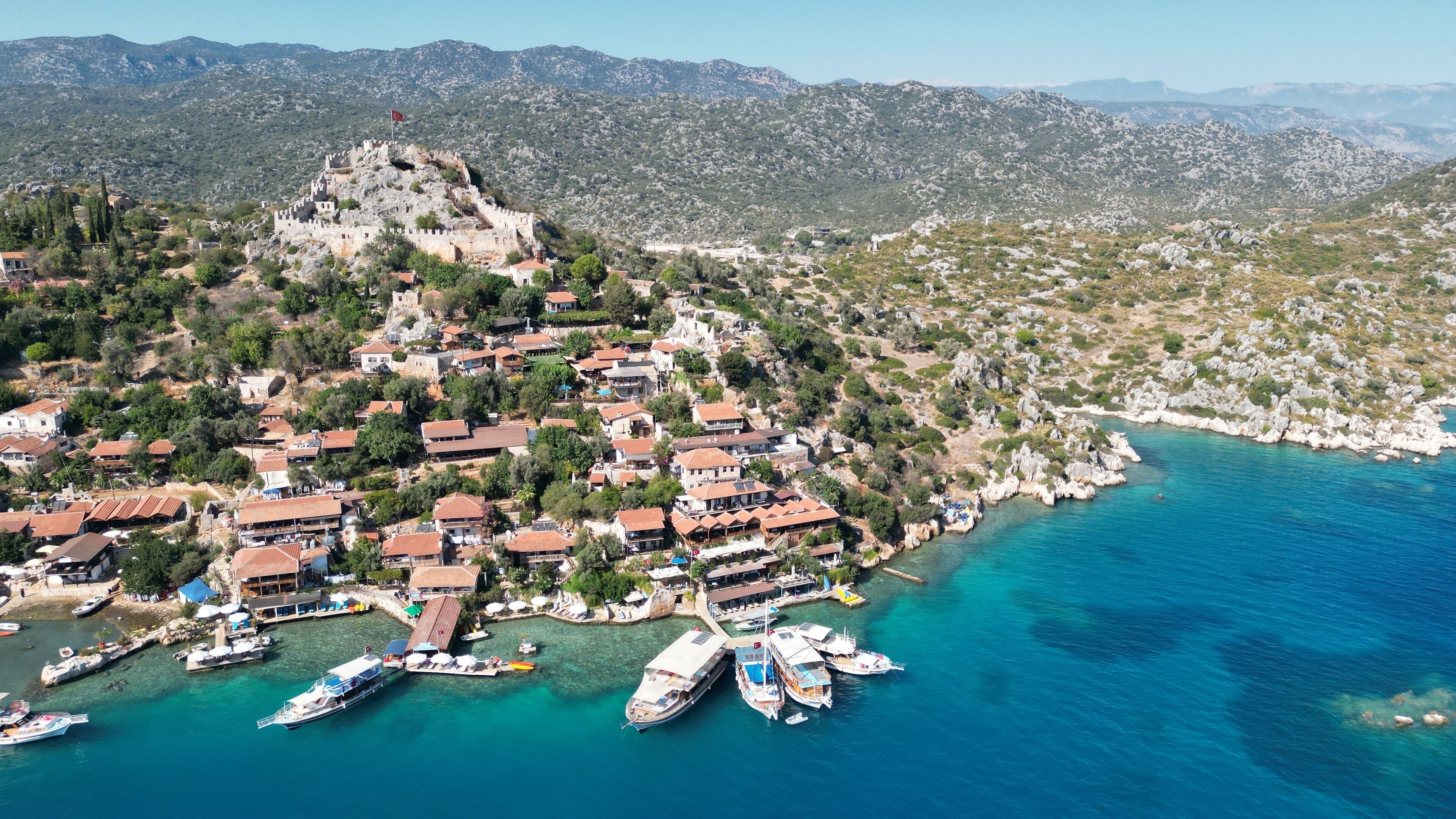 An aerial view of a coastal town with a historic castle on a hill, surrounded by houses and boats docked at the harbor.