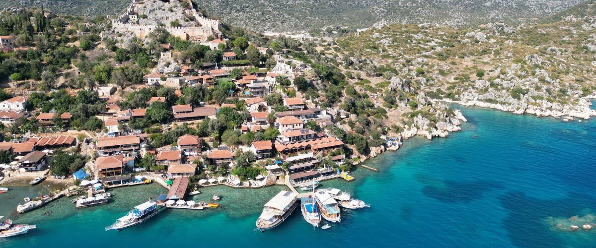 An aerial view of a coastal town with a historic castle on a hill, surrounded by houses and boats docked at the harbor.