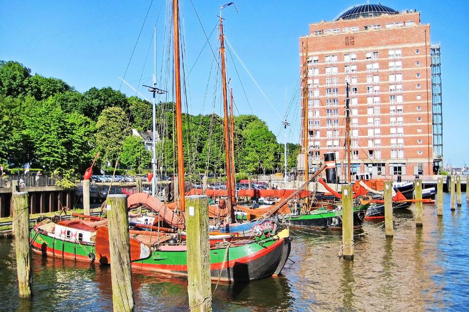 A harbor with colorful boats docked at wooden piers, a tall brick building, and lush green trees in the background.