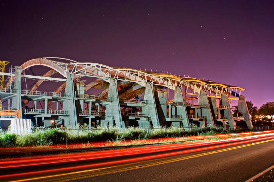 Uma grande estrutura de ponte iluminada à noite, com rastros de luz deixados por veículos em movimento.
