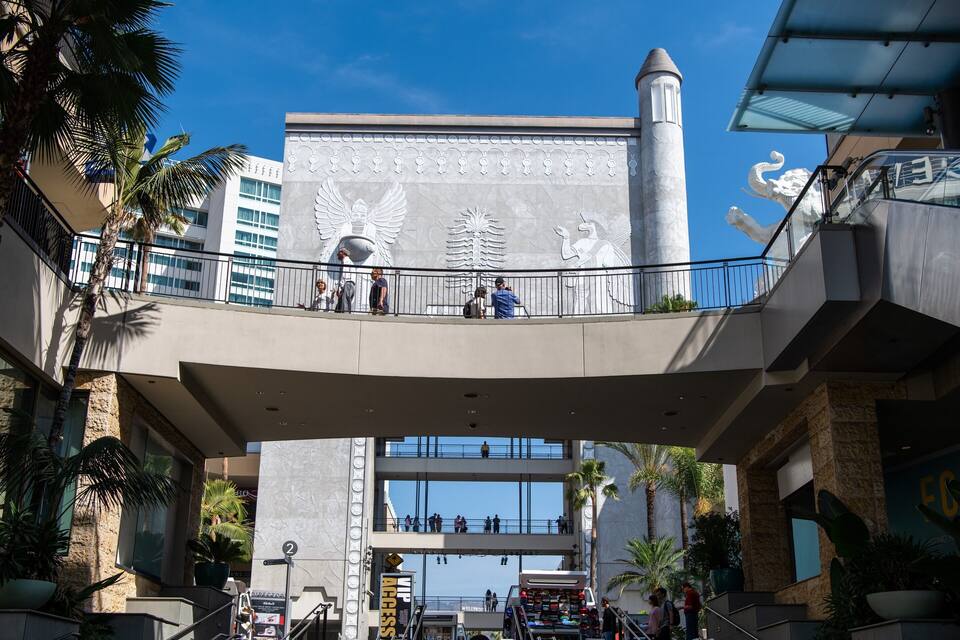 A modern building with a large mural, palm trees, and a pedestrian bridge.