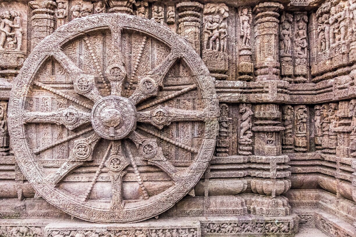 Uma grande roda de pedra ricamente esculpida, com padrões e figuras detalhadas, em frente à arquitetura ornamentada de um templo.