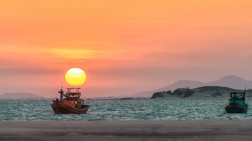A sunset over a calm sea with two boats and distant mountains.