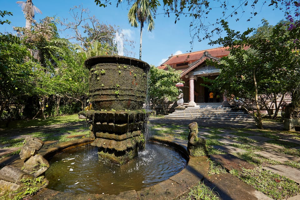 A traditional stone water feature with a cascading waterfall, surrounded by lush greenery and a building with a red roof in the background.