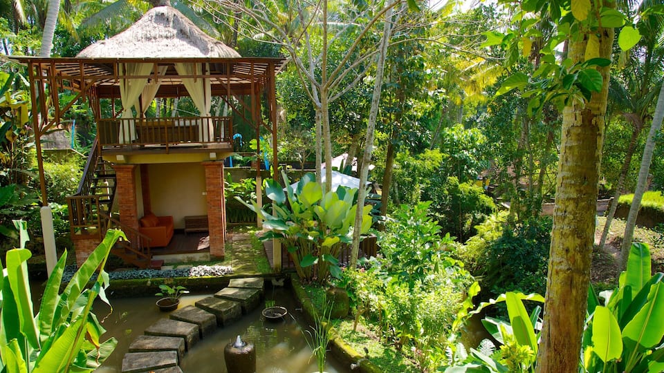 A thatched-roof gazebo with a wooden staircase leading to a balcony, surrounded by lush greenery and a small water feature.