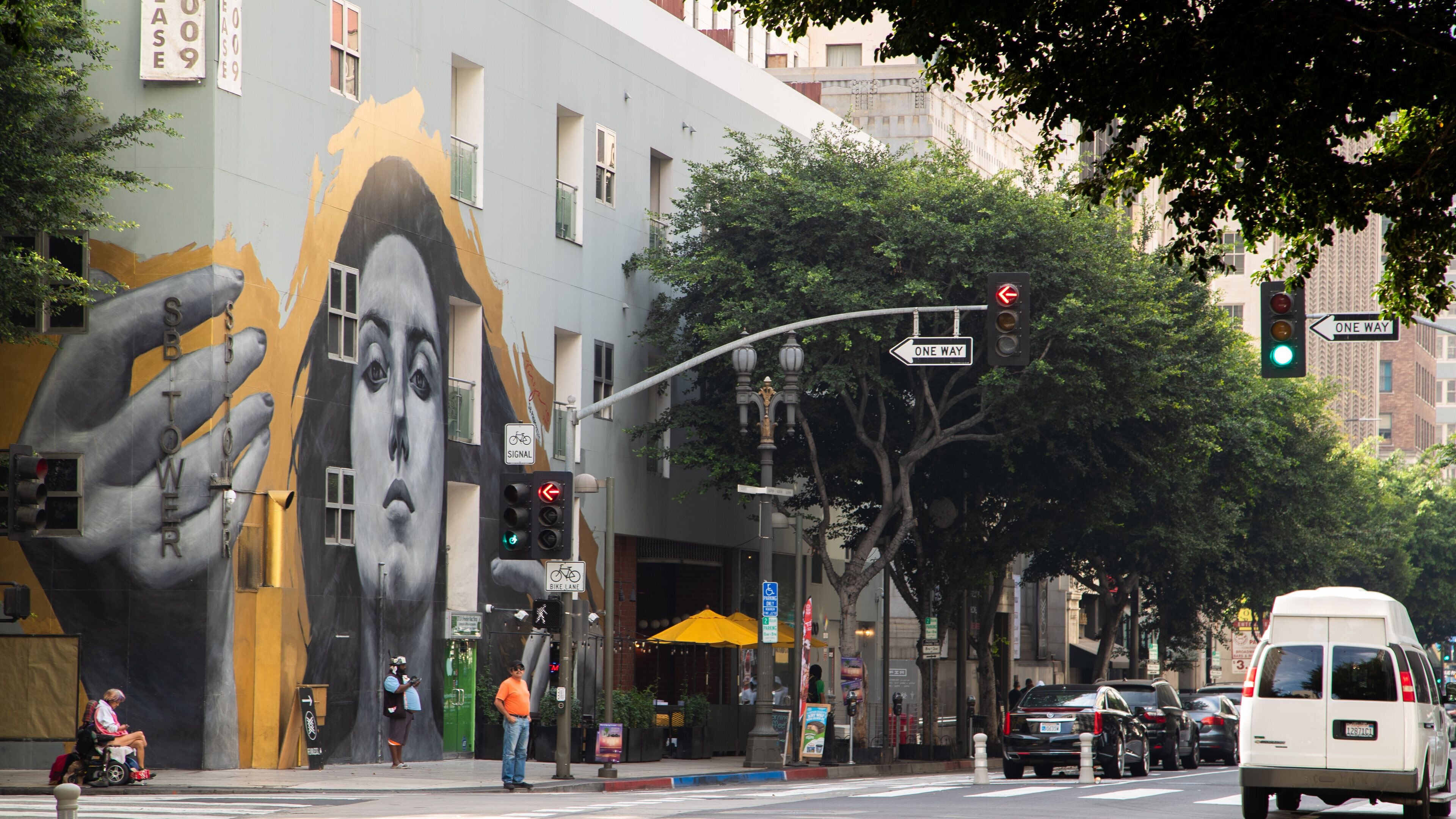 Um mural com o rosto de uma mulher numa fachada, um semáforo, placa de mão única e uma carrinha branca.