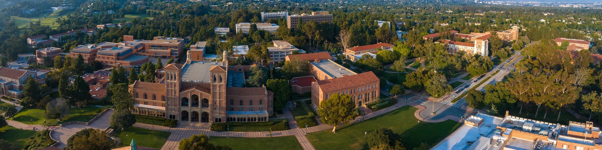 Aerial view of UCLA campus in Westwood, Los Angeles, showcasing Romanesque Revival architecture, modern buildings, green spaces, and a serene city backdrop under a partly cloudy sky.