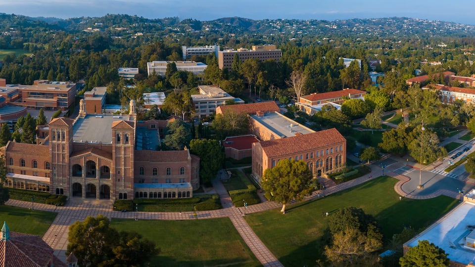 Aerial view of UCLA campus in Westwood, Los Angeles, showcasing Romanesque Revival architecture, modern buildings, green spaces, and a serene city backdrop under a partly cloudy sky.