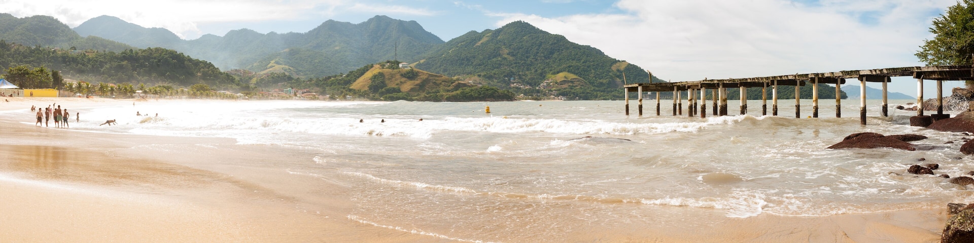 Una playa con olas, un muelle y montañas al fondo.