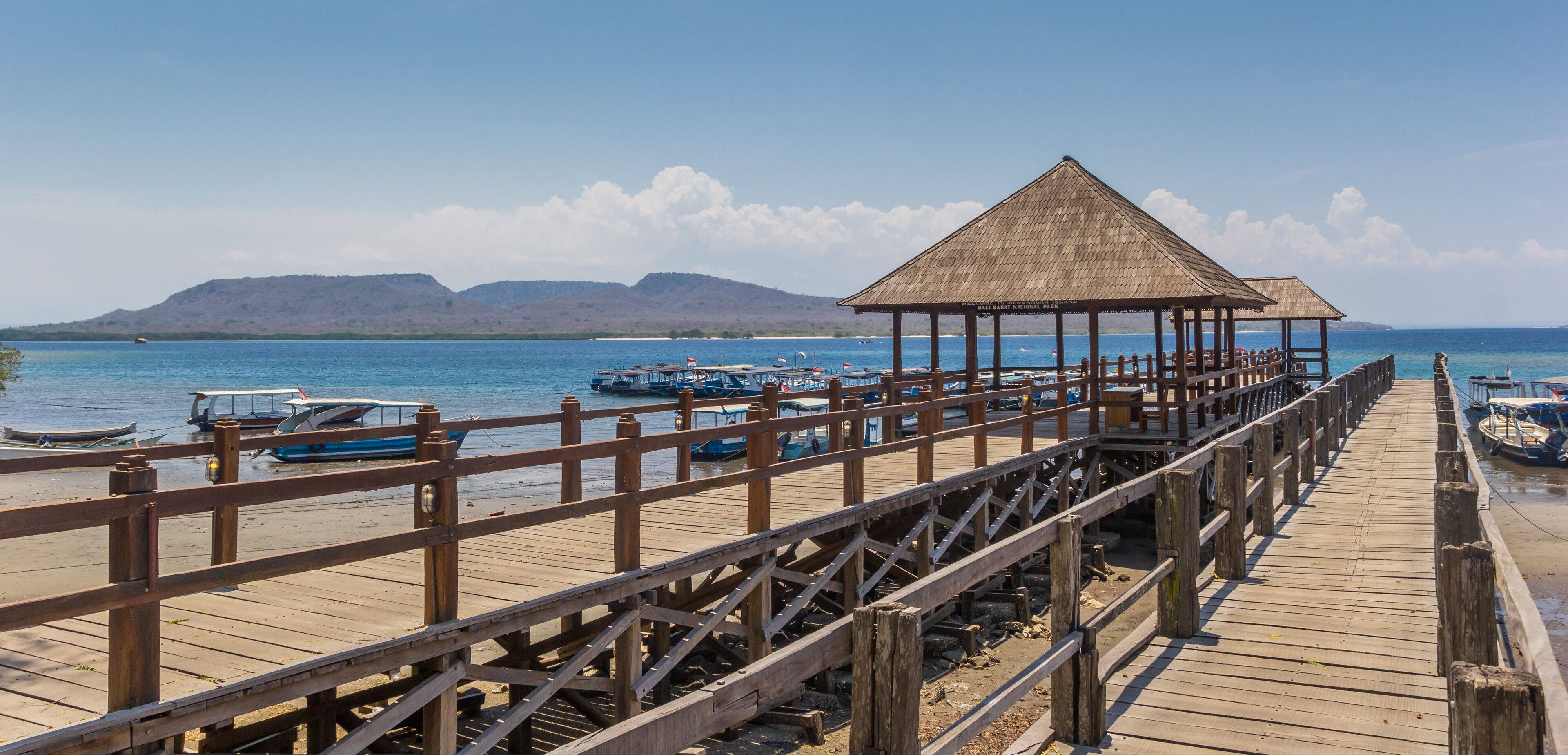 A wooden pier with a thatched-roof gazebo, boats, and a mountainous landscape in the background.