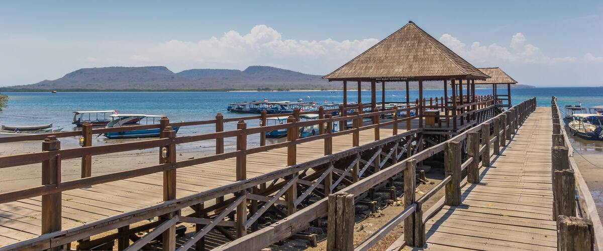 A wooden pier with a thatched-roof gazebo, boats, and a mountainous landscape in the background.