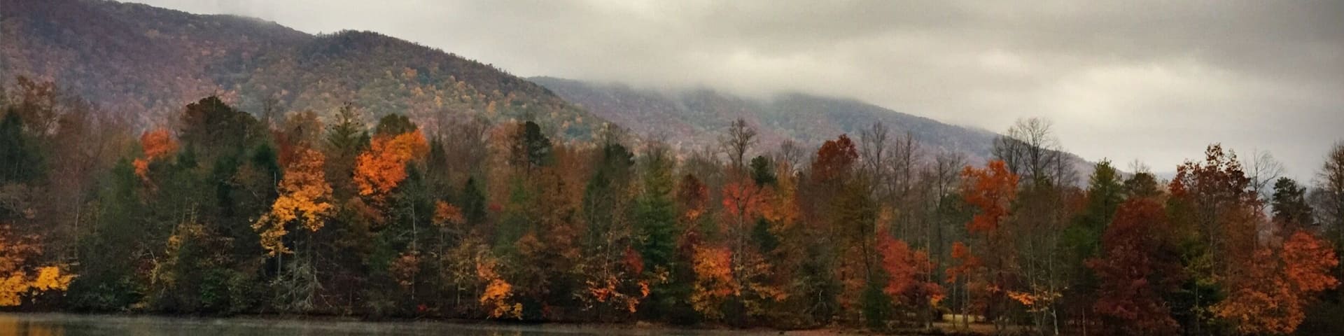 A calm lake reflecting autumn trees with orange and green foliage, surrounded by a misty mountain range.