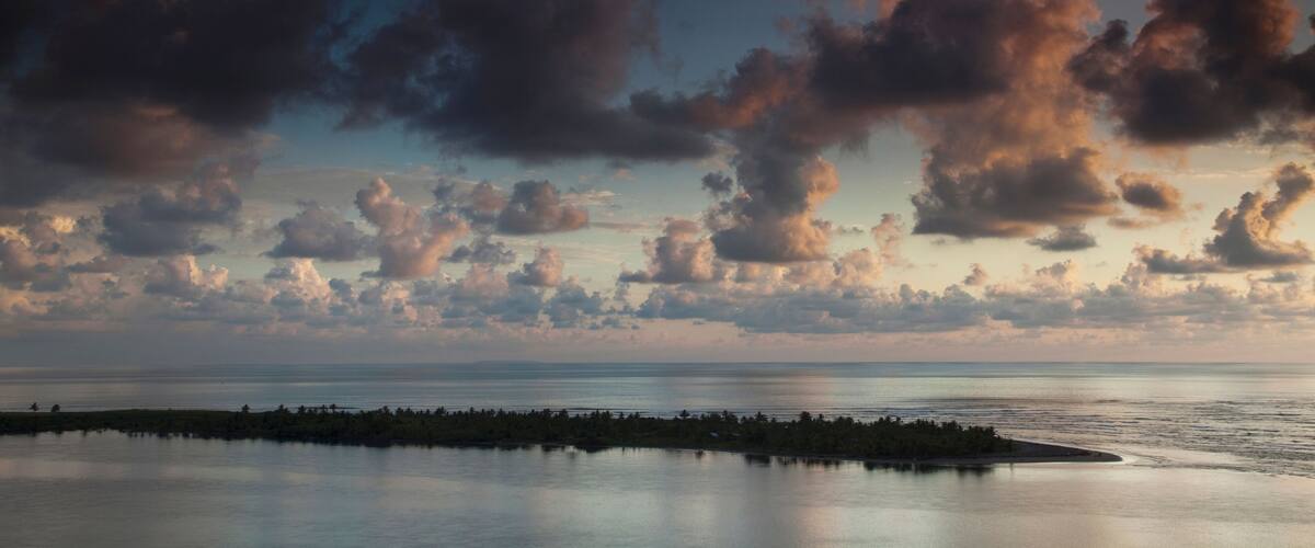 A coastal landscape with a calm sea, a small island with vegetation, and a sky filled with clouds.