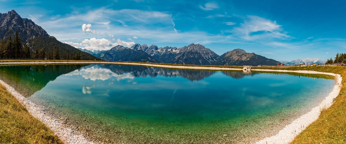 A clear lake reflecting mountains, a gravel path, and a wooden fence.