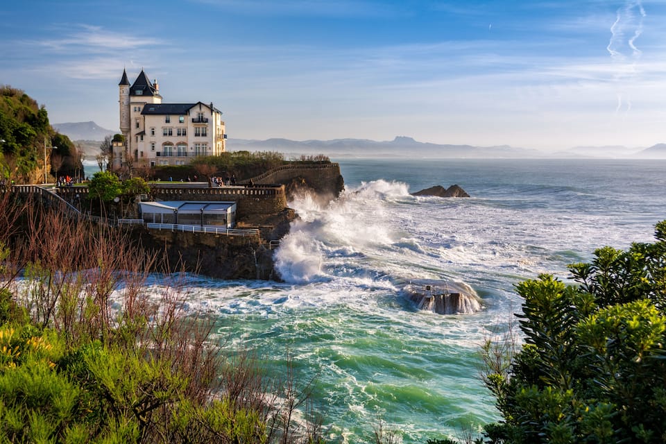A coastal castle overlooking turbulent sea waves, a rocky shoreline, and distant mountains.