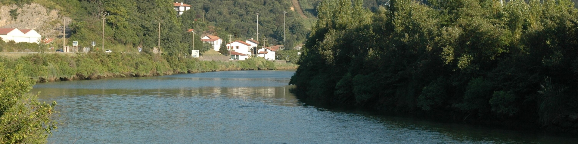 La Bidassoa, vue vers l'amont depuis la D811 près de la redoute Louis XIV. La commune de Biriatou est à gauche (en rive droite) de la Bidassoa (le village lui-même est derrière nous) ; l'Espagne est à droite (en rive gauche). Le hameau d'Aruntz et son pylône sont sur la première hauteur à mi-plan ; ce qui semble être un chemin menant tout droit vers ce sommet, est le passage de lignes à haute tension. La barre rocheuse du Lumaberde est légèrement sur sa gauche (derrière ce sommet), et plus loin derrière le mont du Calvaire (277 m alt.). Le pont de Bénobie, sur le territoire d'Urrugne, est à environ 580 m derrière nous. Le pont de l'autoroute A63 est devant nous, juste après le coude de la rivière.