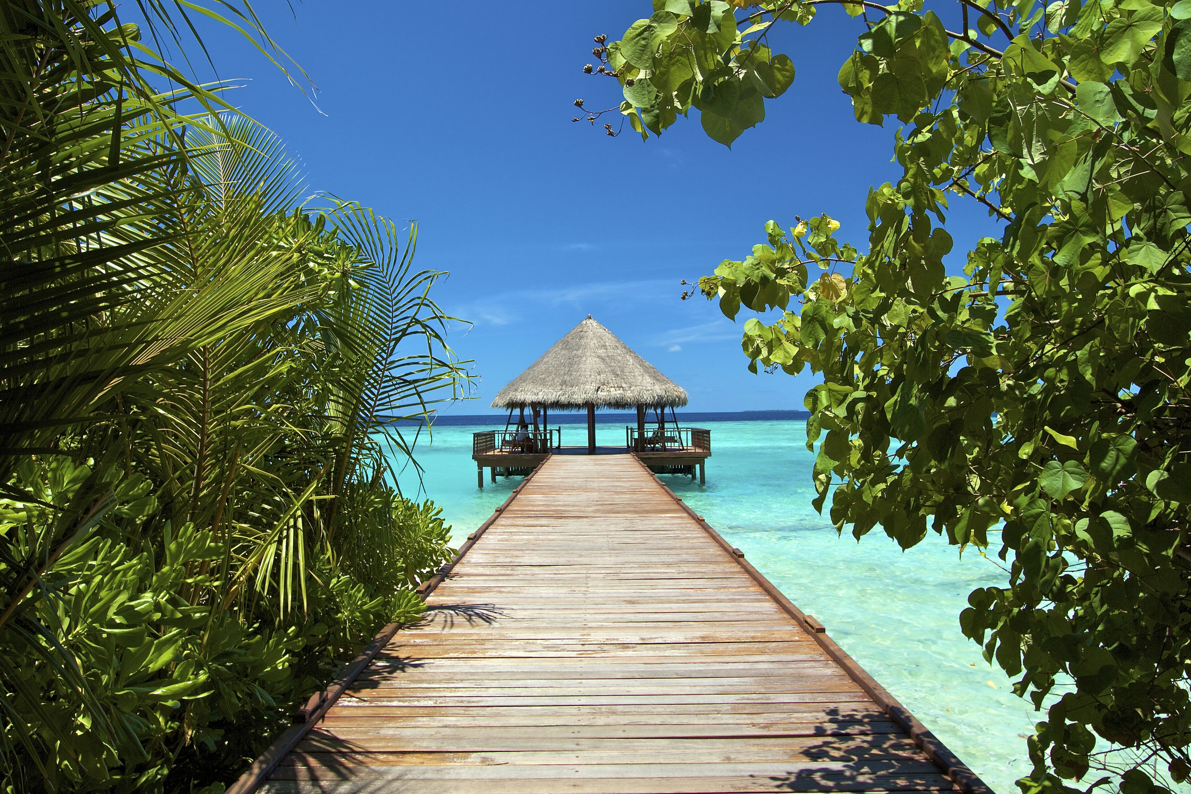 A wooden pier leading to a thatched-over structure on a clear blue sea.