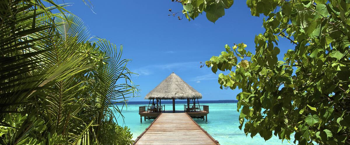 A wooden pier leading to a thatched-over structure on a clear blue sea.