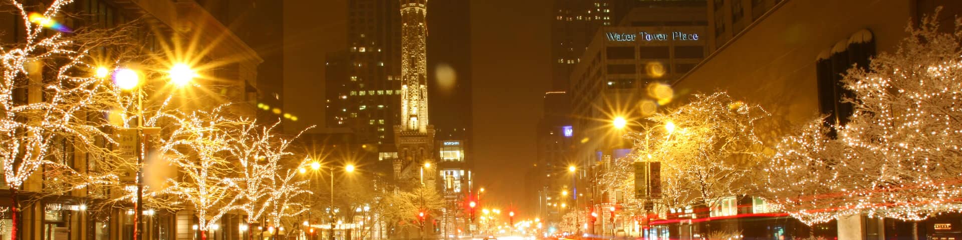 A city street at night with illuminated buildings, a snow-covered sidewalk, and light trails from moving vehicles.