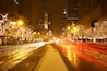 A city street at night with illuminated buildings, a snow-covered sidewalk, and light trails from moving vehicles.