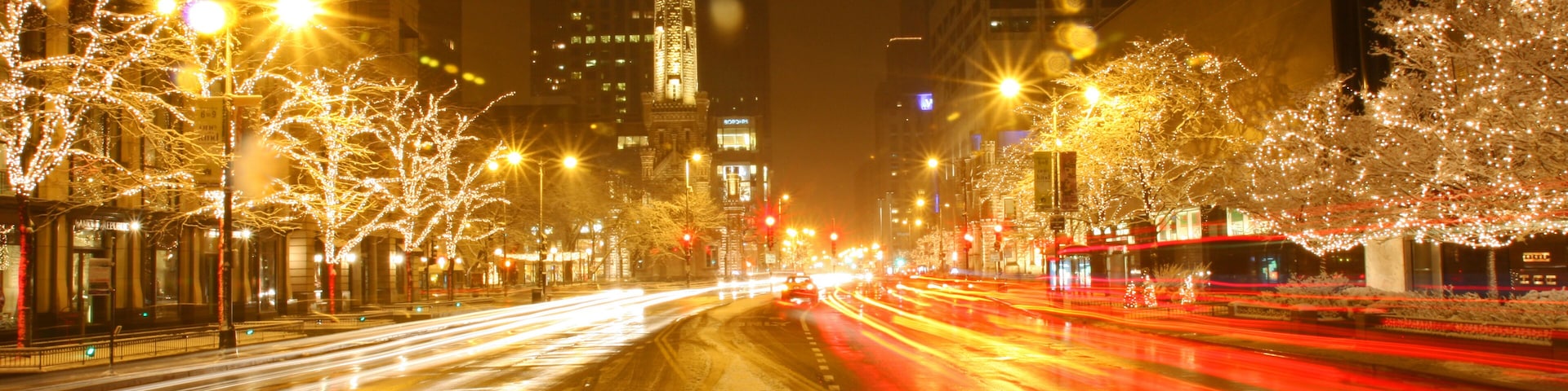 Rua urbana à noite com edifícios iluminados, calçada coberta de neve e rastros de luz de veículos em movimento.