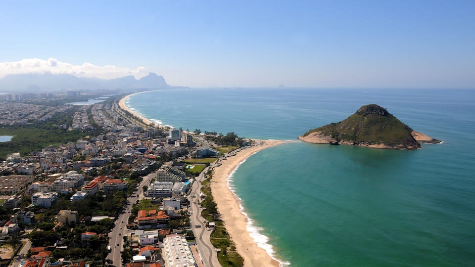 Paisagem costeira com uma praia em destaque, uma colina grande e uma cadeia de montanhas ao longe.