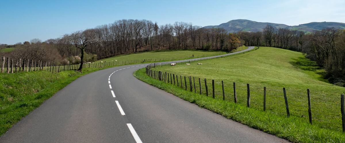 A curving country road flanked by green fields and a fence, with hills in the background.