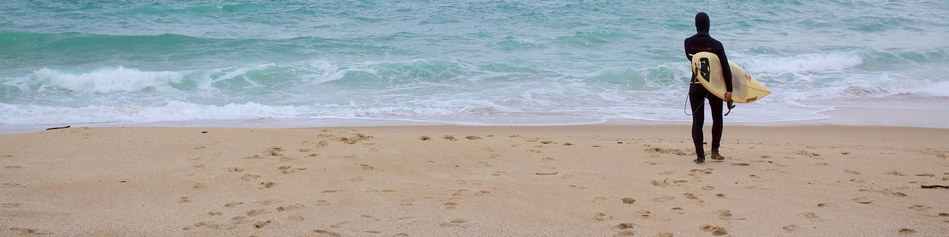 A person in a wetsuit walking on a sandy beach with footprints, holding a surfboard, and looking out at the ocean under a cloudy sky.