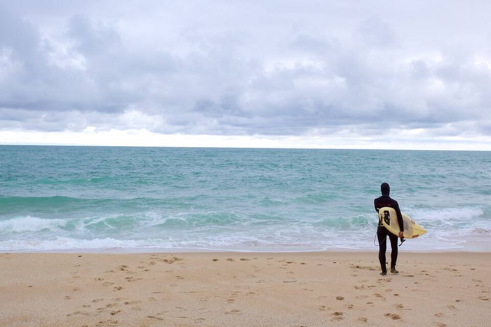 A person in a wetsuit walking on a sandy beach with footprints, holding a surfboard, and looking out at the ocean under a cloudy sky.