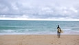 Une personne vêtue d’une combinaison de plongée marche sur une plage de sable, laissant des empreintes de pas derrière elle. Elle tient une planche de surf et regarde l’océan sous un ciel nuageux.