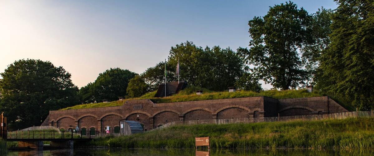A brick structure with arched windows and a small building with a green roof, reflecting in a calm body of water.