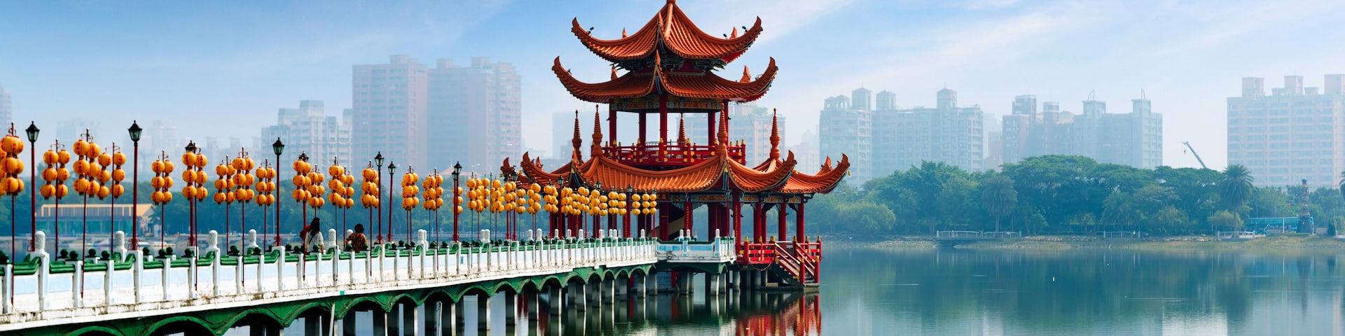 Um pavilhão tradicional chinês sobre um píer, acima de águas calmas, com um horizonte urbano ao fundo.