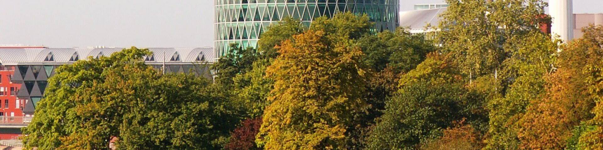 A modern cylindrical building with a geometric glass facade, surrounded by a park with trees and a pathway.