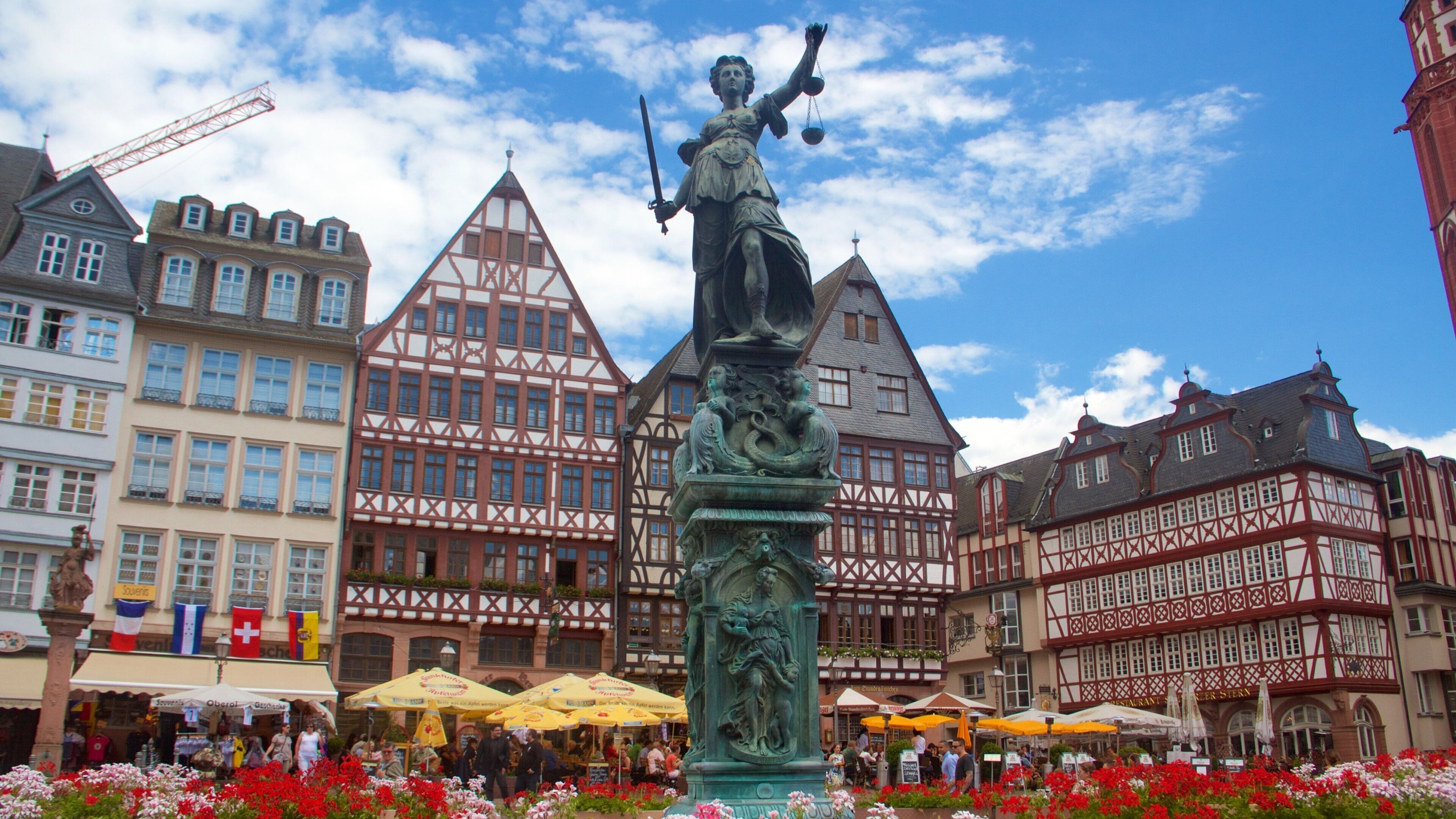 A statue of a woman holding a sword and a balance scale, surrounded by colorful flowers, with traditional half-timbered buildings in the background.