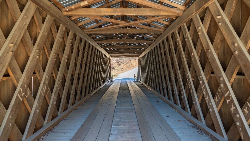 A wooden pedestrian bridge with a metal roof, featuring a symmetrical design and numbered beams.