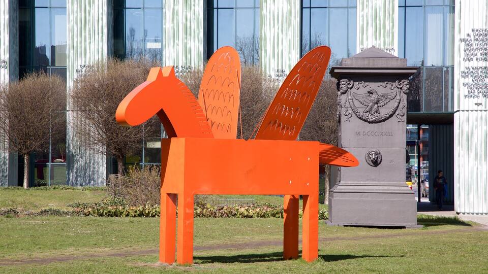 A large orange sculpture of a horse in a grassy area with a modern building and a monument in the background.