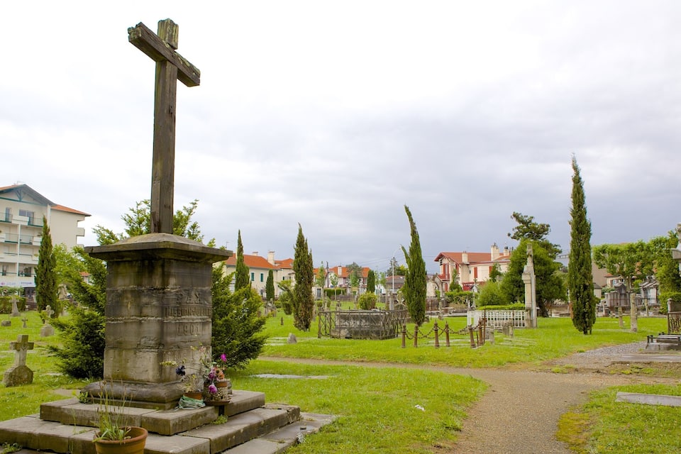 Un cementerio con una cruz de madera destacada, monumentos de piedra y un sendero.