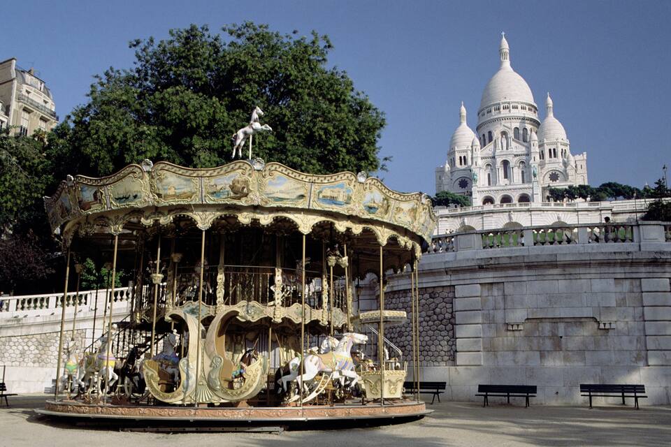 A carousel with ornate decorations and horses, set against a backdrop of a large white domed building and lush green trees.