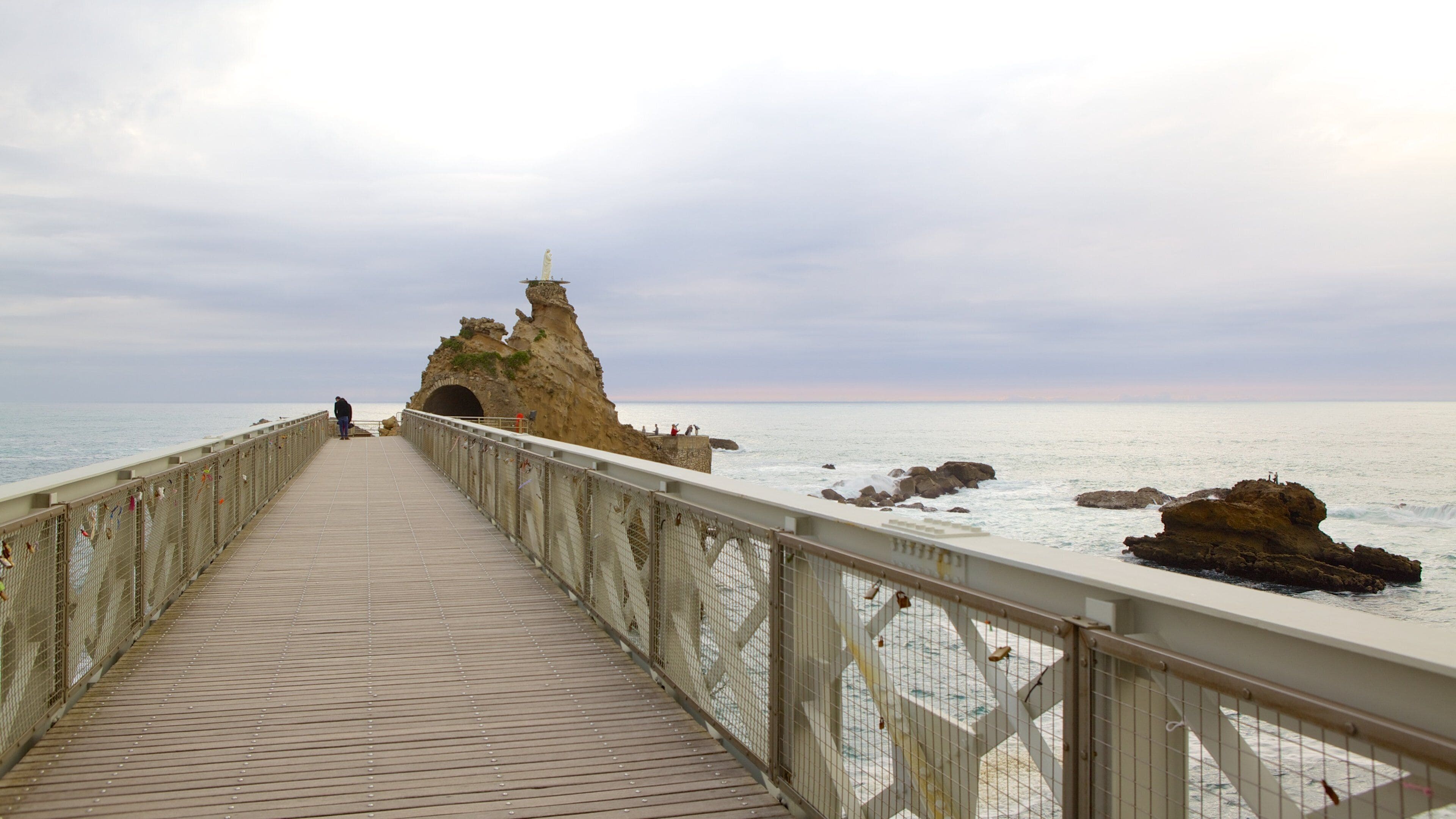 A wooden pier leading to a rocky coastline with a statue at the end.
