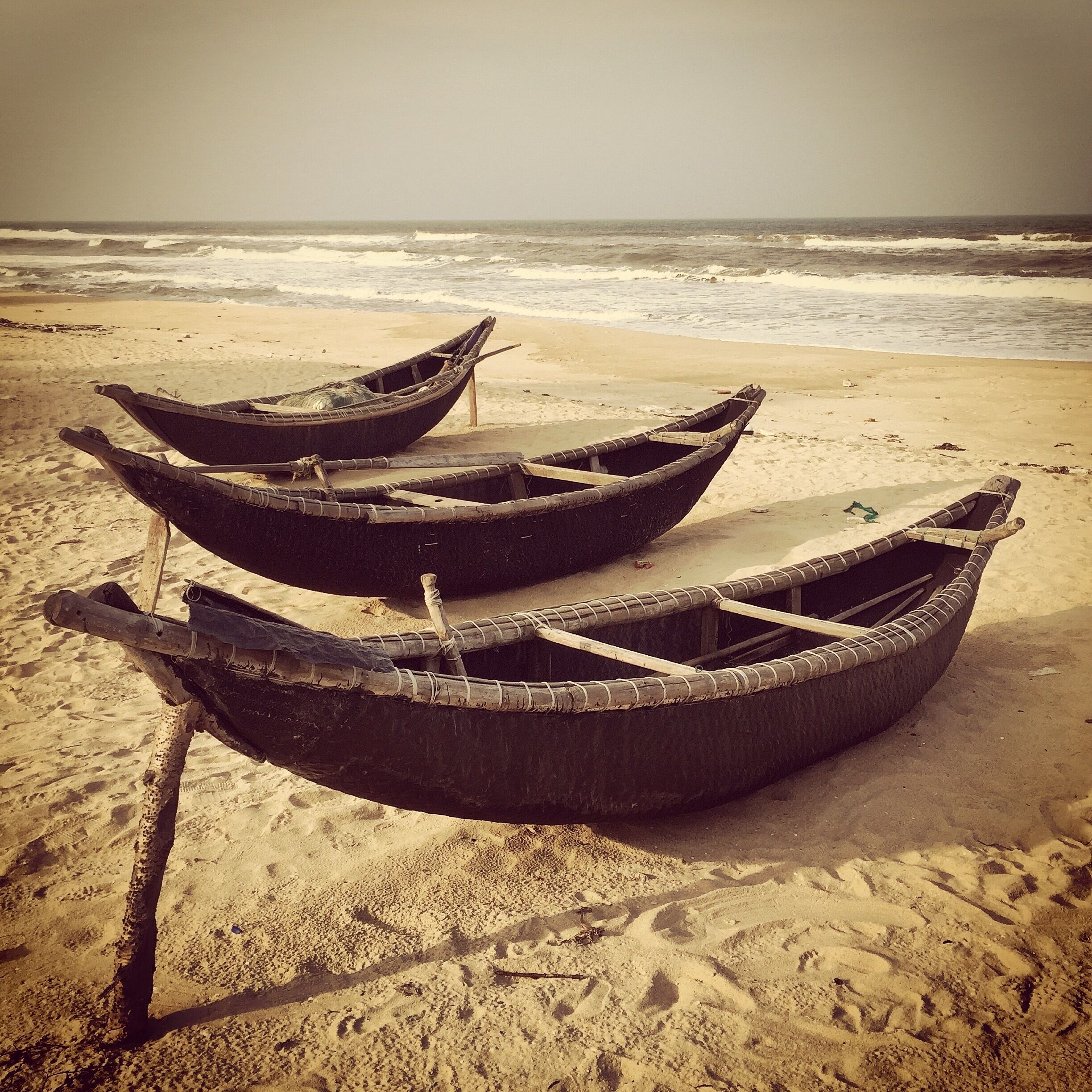 Three traditional boats on a sandy beach with the ocean in the background.