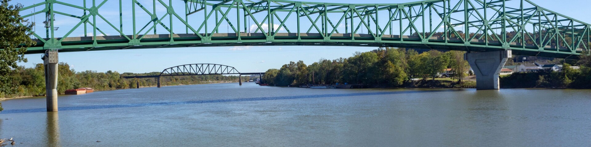 Uma ponte metálica verde sobre um rio, com uma ponte menor ao fundo.