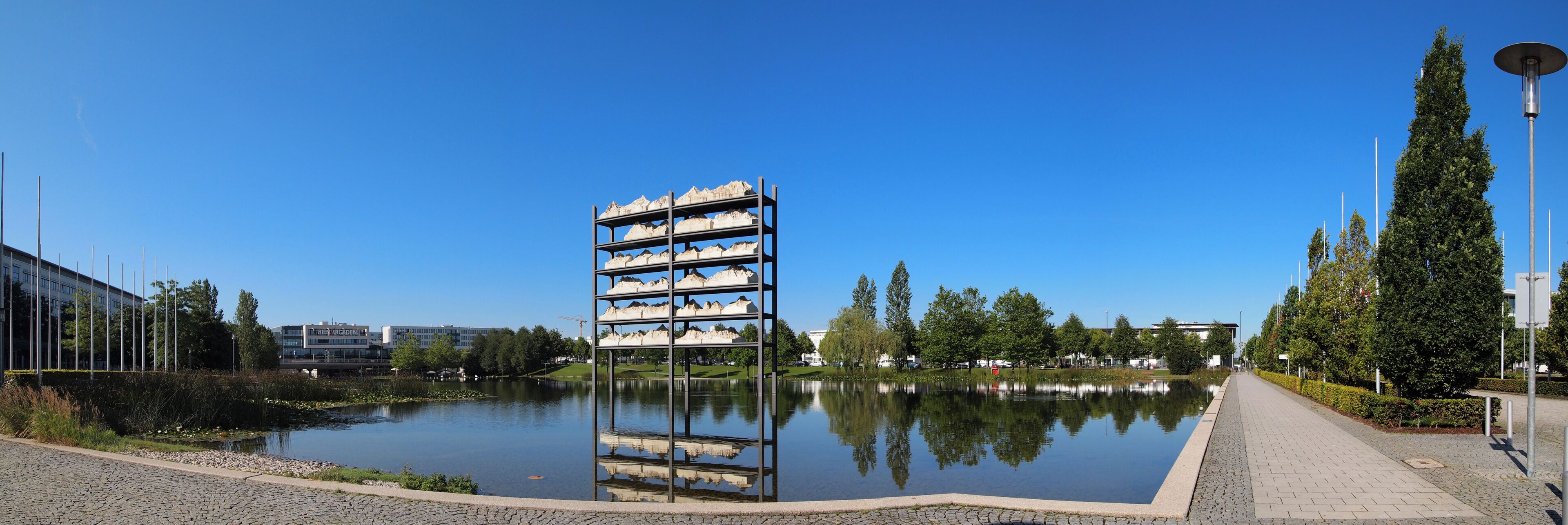 A calm lake with a central sculpture, surrounded by trees and a paved walkway.