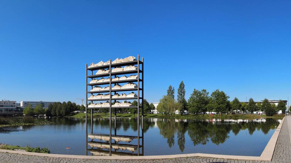 A calm lake with a central sculpture, surrounded by trees and a paved walkway.