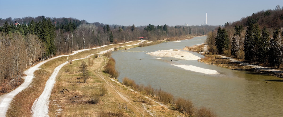 A river with a dirt path alongside it, surrounded by trees and a distant industrial facility.